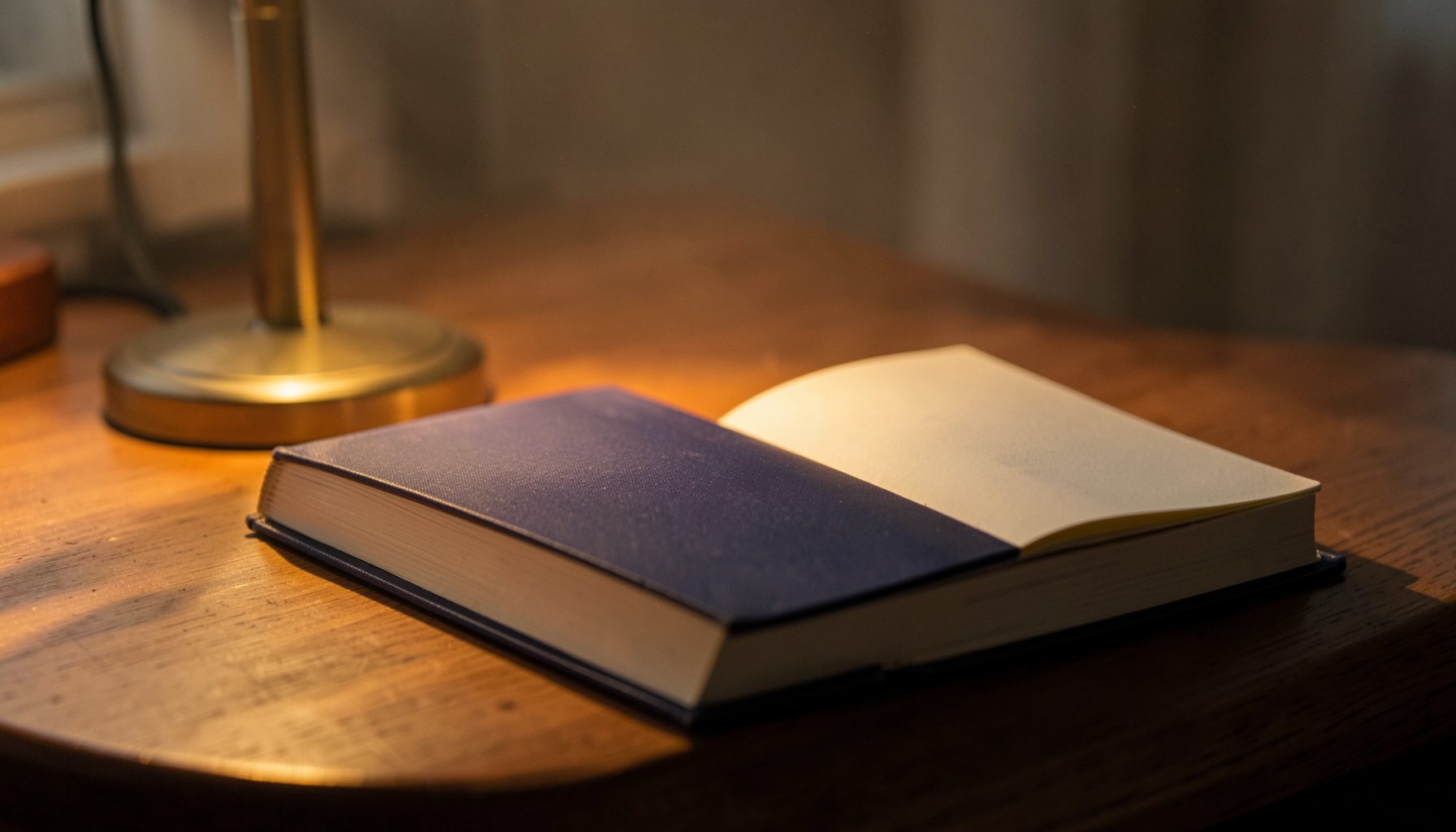 A hardcover book resting on a wooden desk beside a reading lamp, warm amber light spilling across the open pages, deep navy and cream tones.