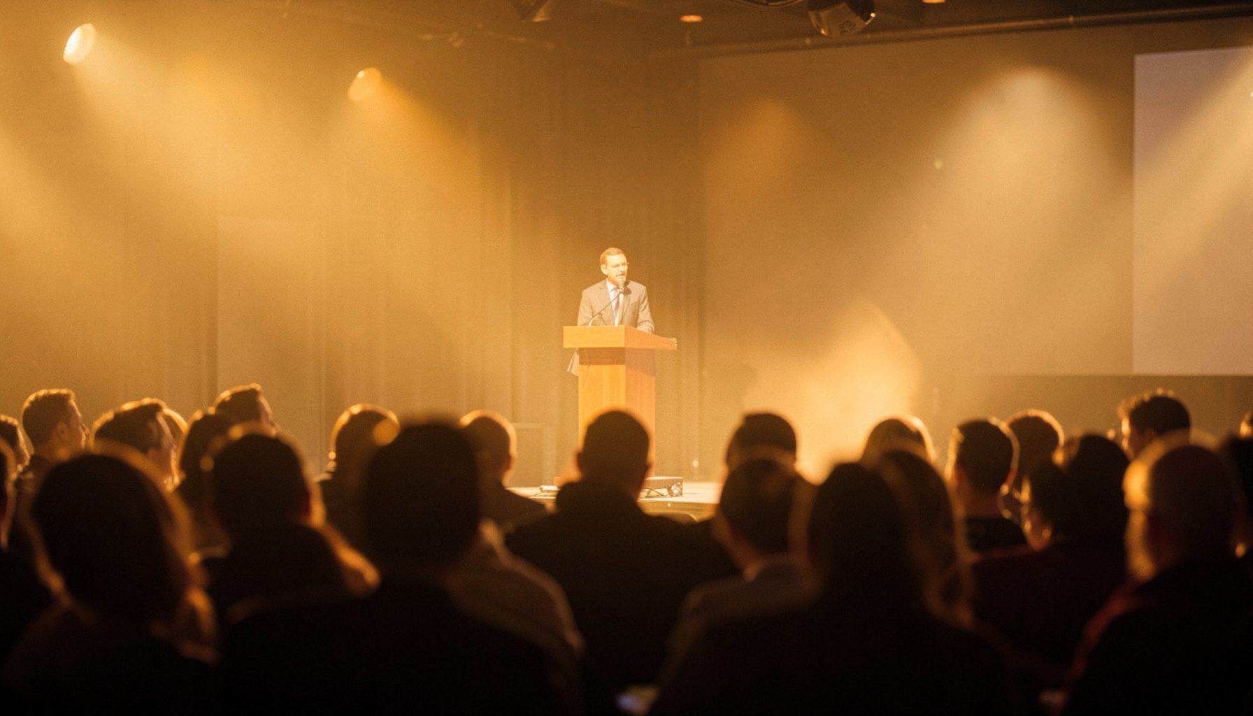 A warmly lit conference stage with a speaker at a podium, audience silhouettes in soft focus, golden and amber tones filling the room