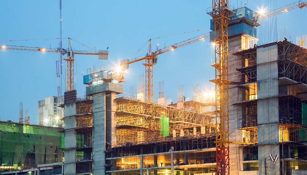 A large multi-story building under construction at dusk or night, with several tall cranes, scaffolding, and concrete pillars silhouetted against a deep blue twilight sky, conveying steady progress and thoughtful structure.